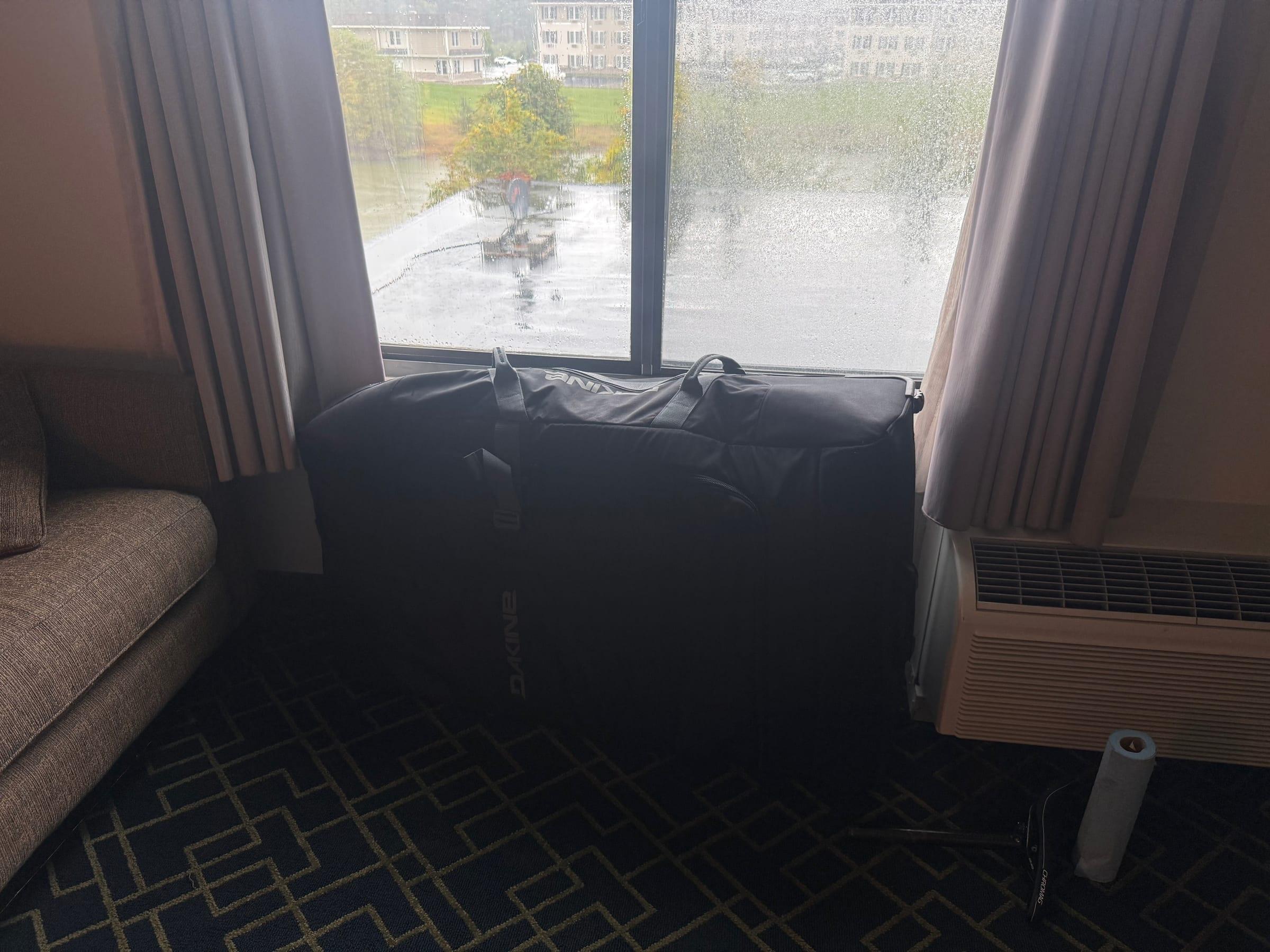 A black Dakine bike bag standing upright in front of a rain-streaked hotel window, with a roll of paper towels and a seatpost on the carpet beside it