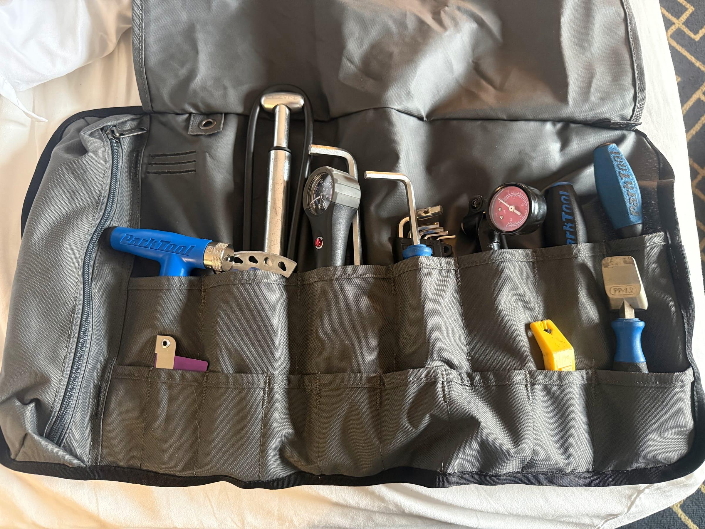 A gray tool roll unrolled on a hotel bed, with Park Tool wrenches and a tire lever, hex keys, a pump, a shock pump gauge, a tire pressure gauge, and other bike tools in its pockets