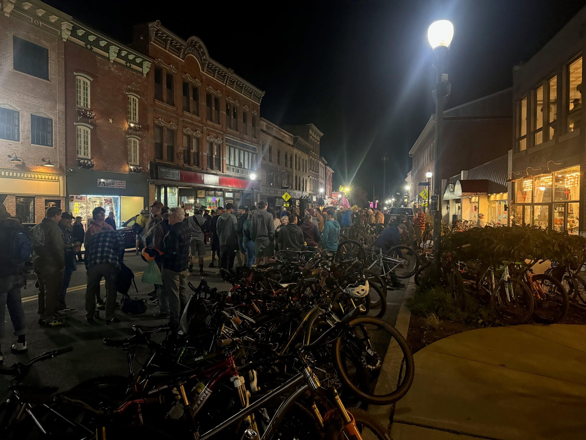 A crowd gathered on a closed downtown street at night in front of brick storefronts, with a large rack of parked mountain bikes in the foreground