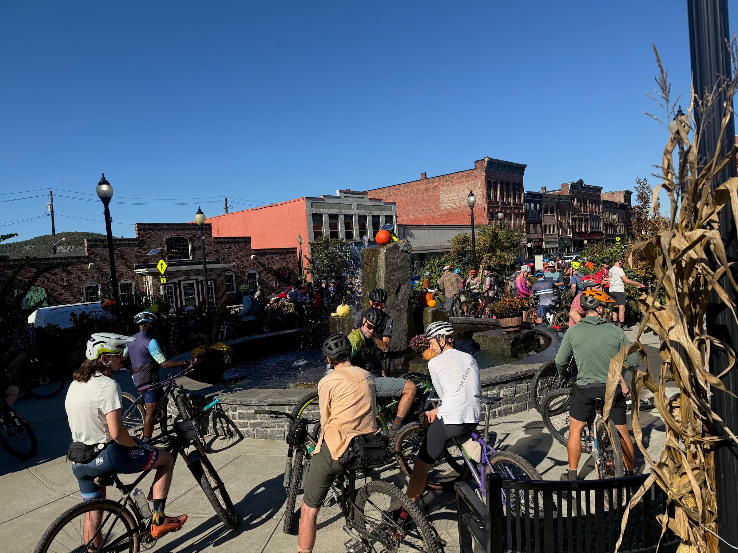 A crowd of riders in helmets standing and sitting on bikes around a downtown fountain on a sunny morning, with brick buildings behind them