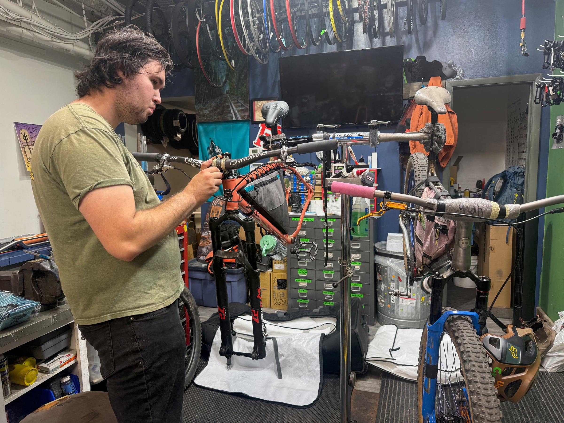 A mechanic at NeverLost Adventures in Port Jervis assembling an orange Surly Karate Monkey on a repair stand, with another bike on the next stand and wheels hanging from the ceiling