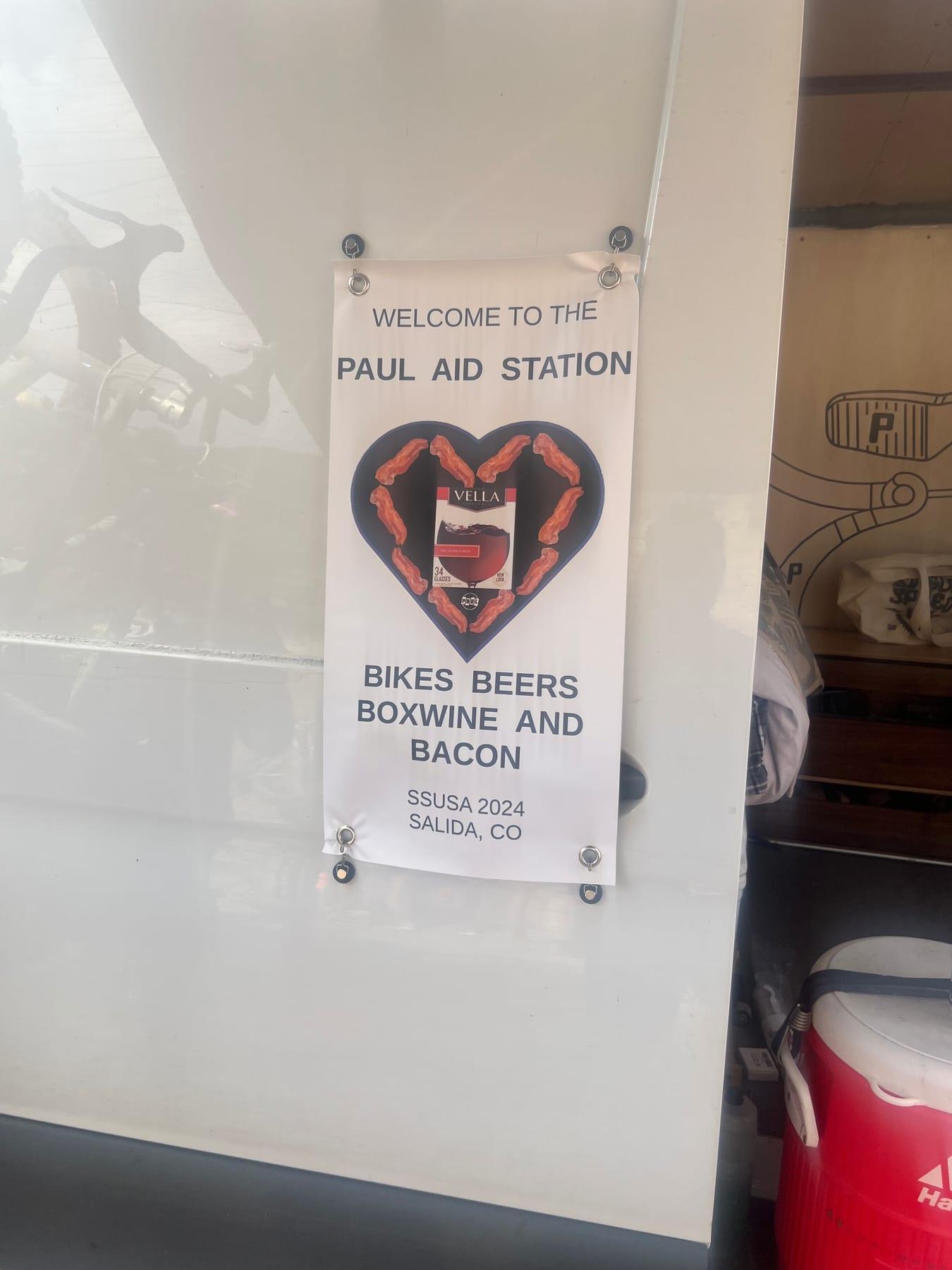 Hand-lettered sign reading WELCOME TO THE PAUL AID STATION, BIKES BEERS BOXWINE AND BACON