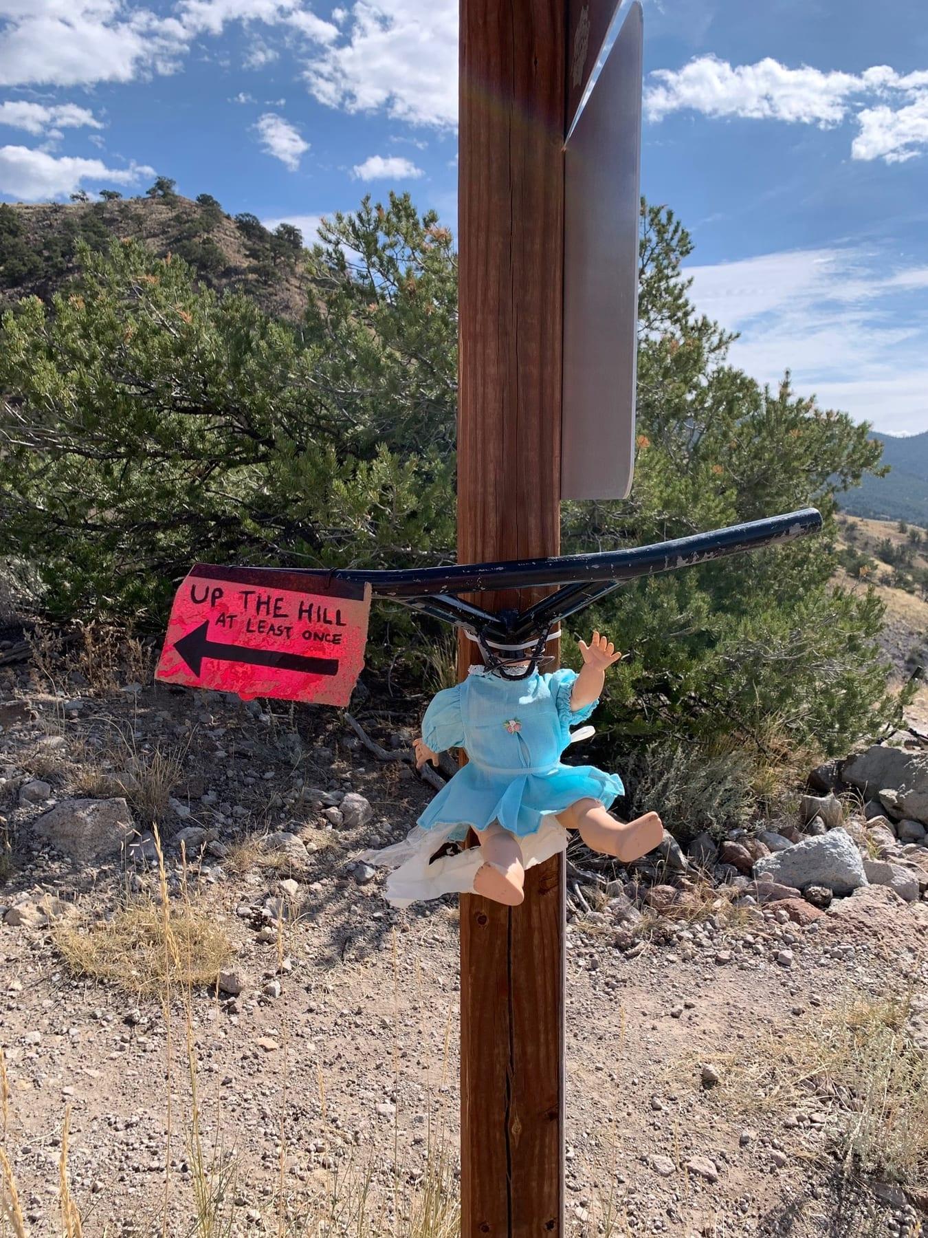 Trailside sign reading OFF THE HILL with a small doll in a tutu attached to the post