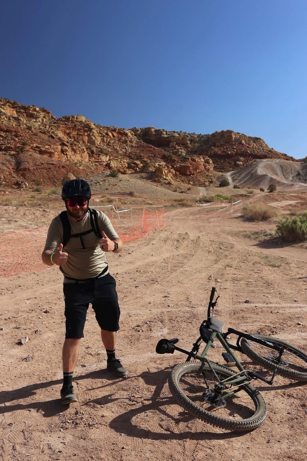 Rider standing over a bike laid down on the trail, desert hills in the background
