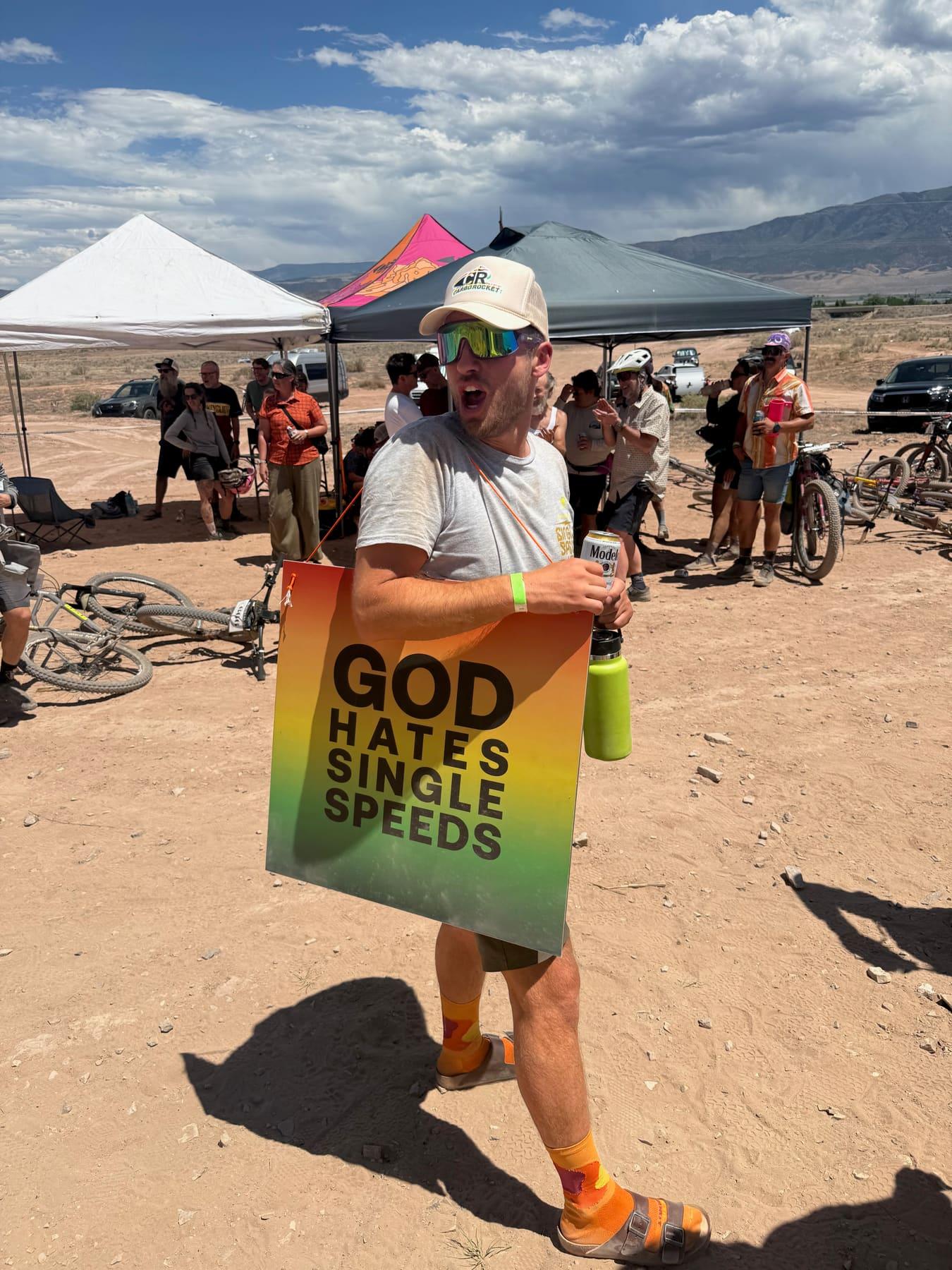 Rider carrying a GOD HATES SINGLE SPEEDS sign and a beer at the Single Speed USA aid station tents in the desert