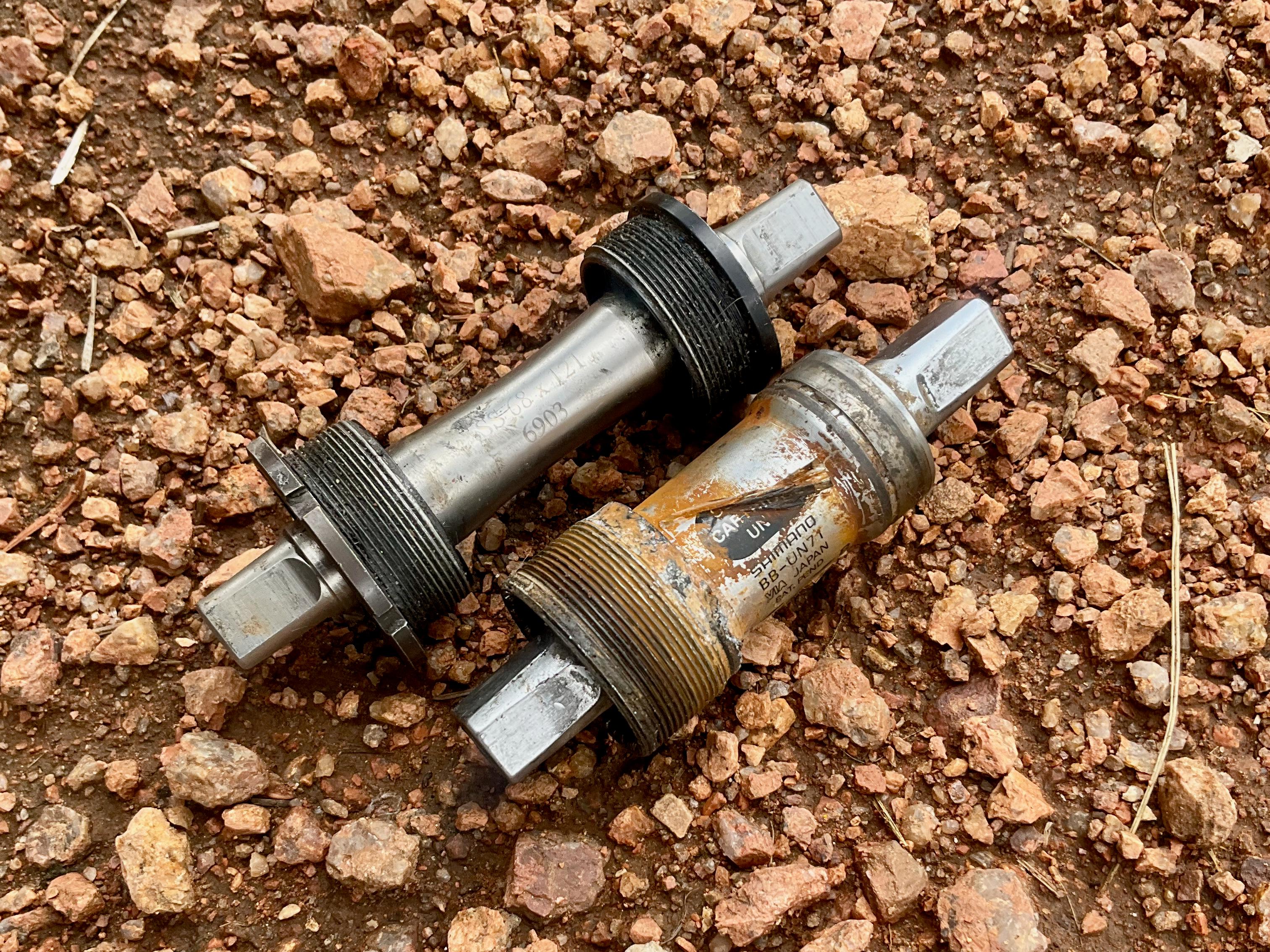 Two old Shimano square taper bottom bracket spindles lying side by side on rocky dirt, one bare silver, one rusted and labeled Shimano BB-UN71