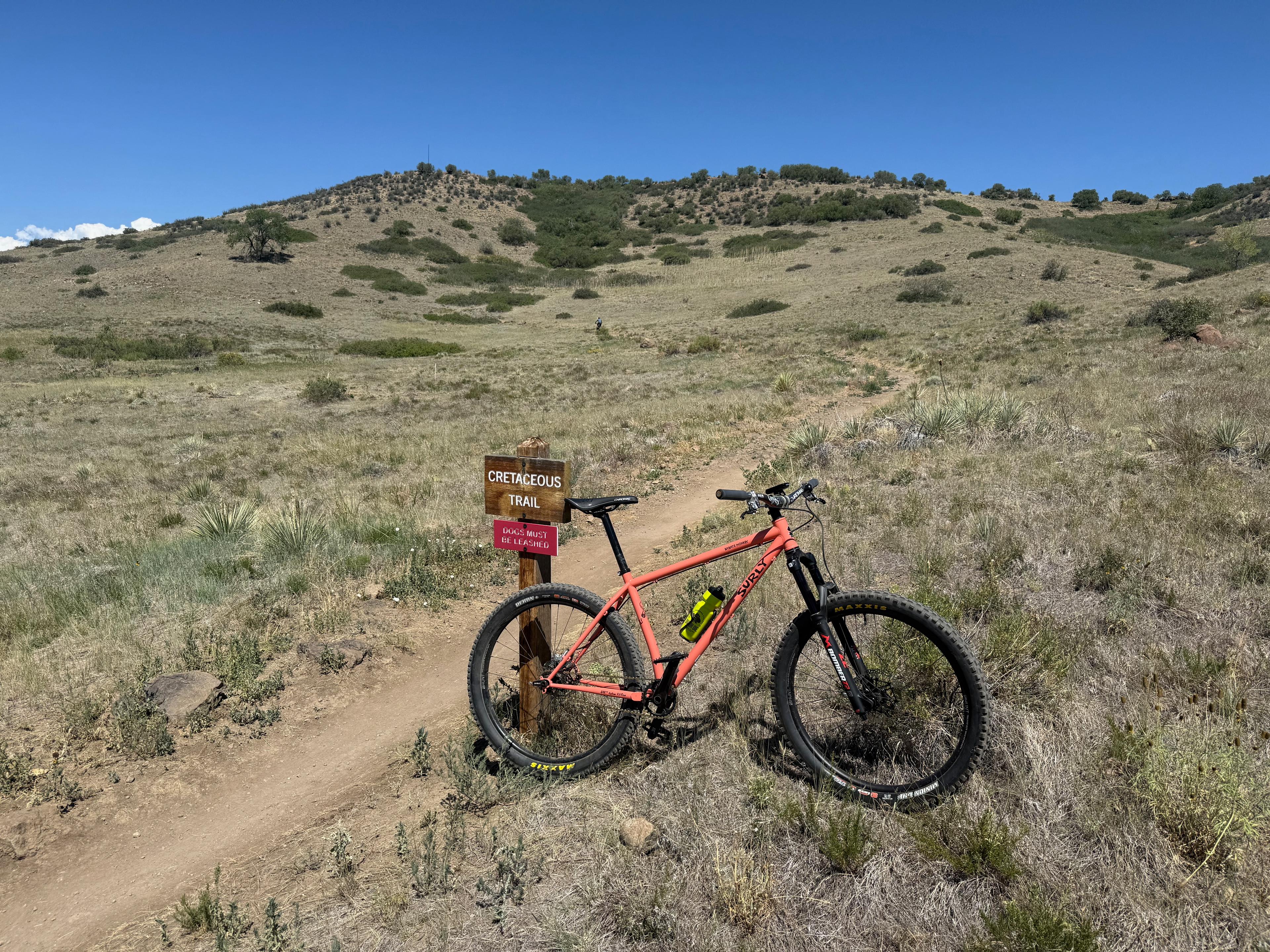 Single-speed mountain bike beside the Cretaceous Trail sign partway up the climb, with a leashed-dogs notice posted below