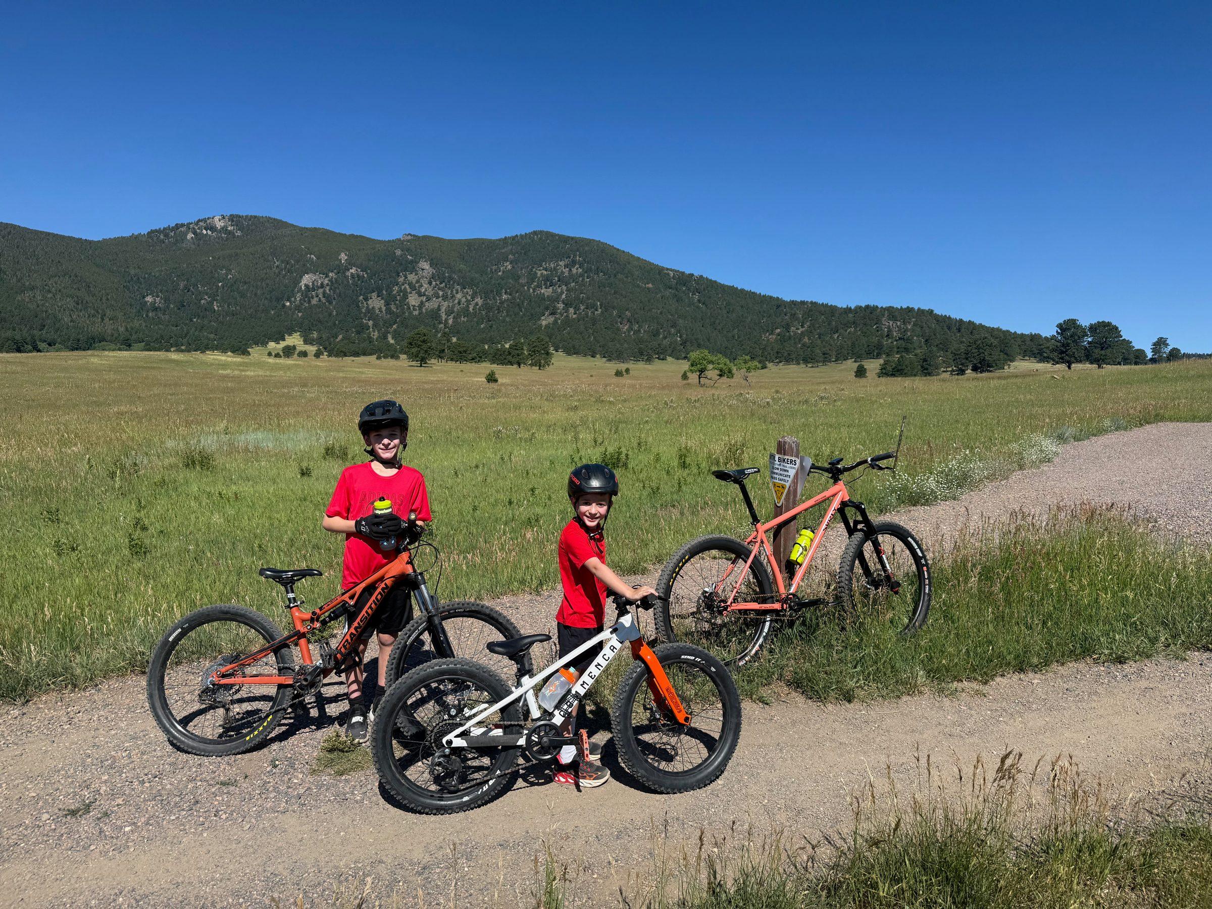 Two boys in red shirts standing with three mountain bikes at a gravel trail junction in a broad grassy meadow, a forested mountain rising behind them