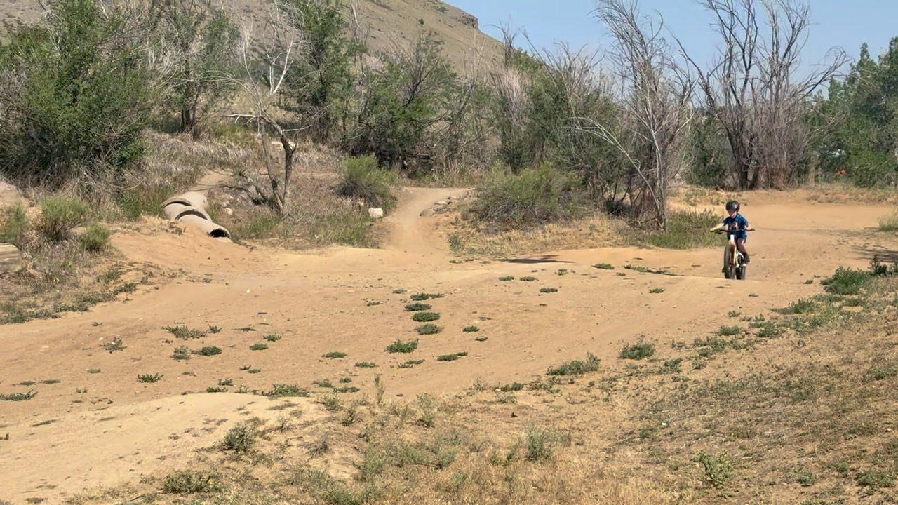 A small child riding a bike across a sandy dirt pump track lined with dry brush, low foothills rising in the background