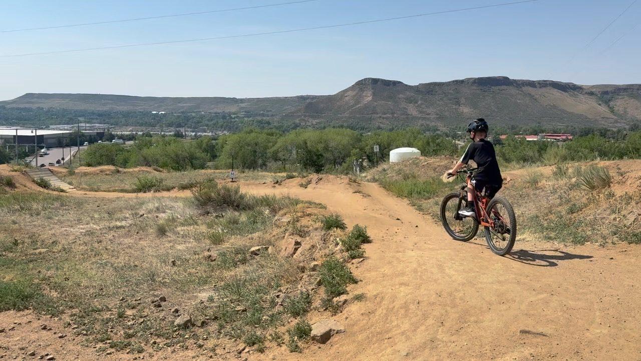 A child on an orange mountain bike riding away down a dirt trail toward a flat-topped mesa on the Front Range, with a town spread out in the valley below