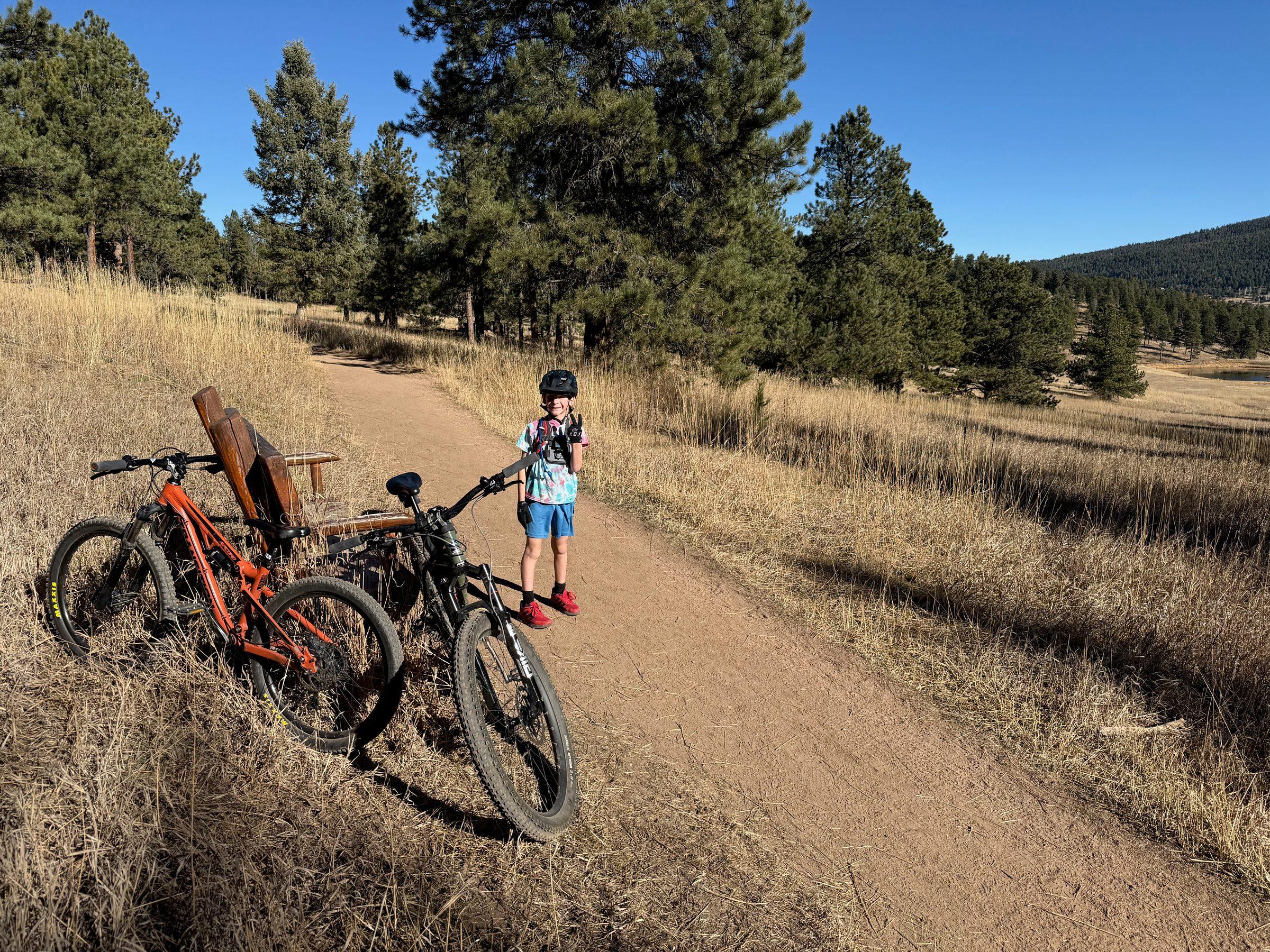 A young child in a helmet and tie-dye shirt standing on a dirt trail beside two mountain bikes, dry grass and ponderosa pines around them under a blue sky